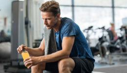 A Young Man Sitting on a Gym Bench Holding a Yellow Sports Drink in His Hands Testosterone Replacement Therapy Near Me